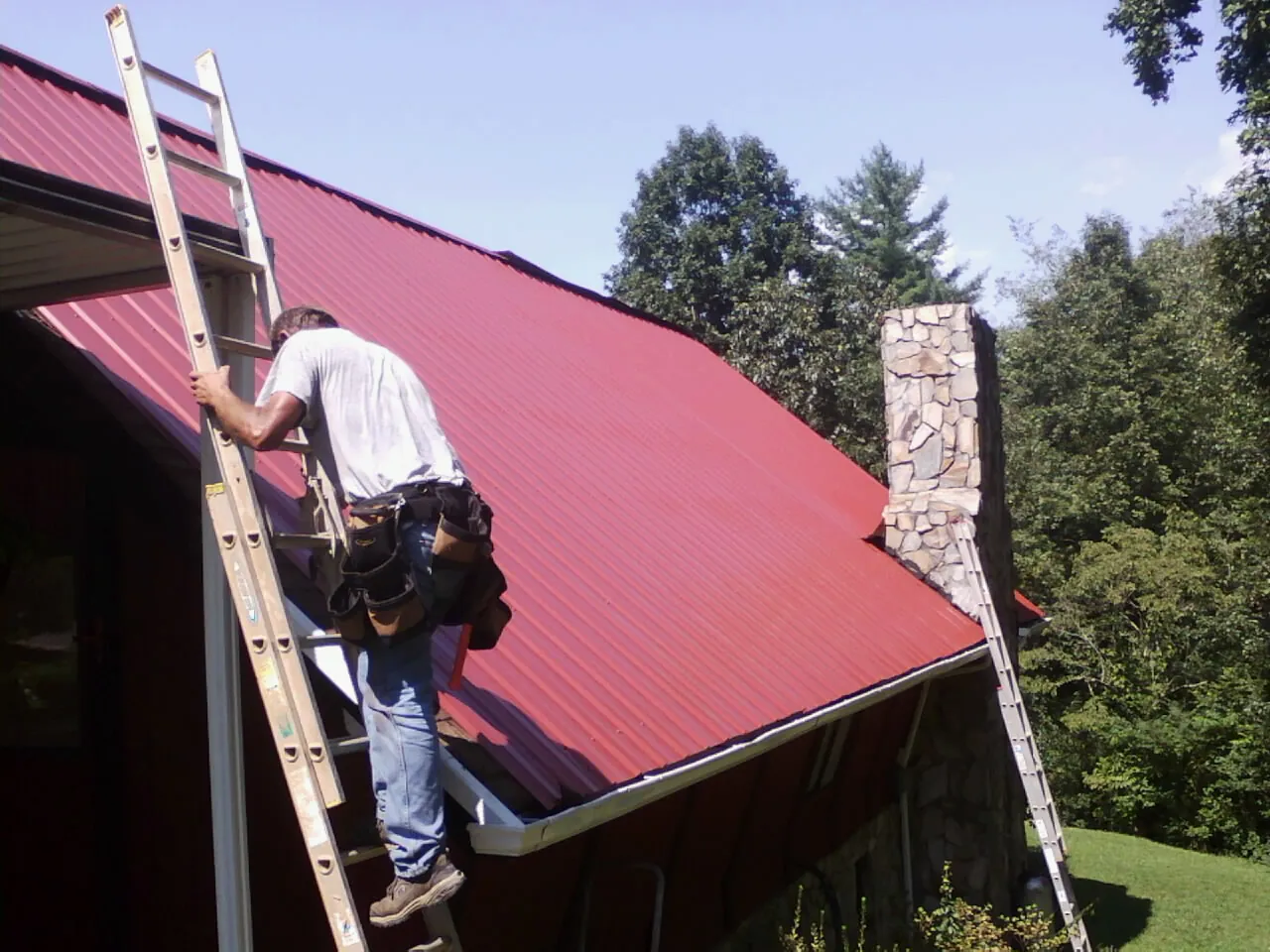 Roofer on a ladder beside a red metal roof and stone chimney — Soffit & Fascia Repair by Roofing Brothers in Virginia